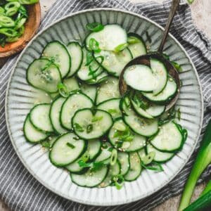 Overhead close-up of an asian cucumber salad recipe in a ribbed bowl, garnished with sesame seeds and sliced scallions, with a spoon lifting a portion