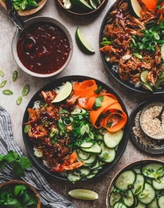 Asian style pulled pork rice bowl surrounded by cucumber salad, lime, cilantro, sesame seeds and extra sauce