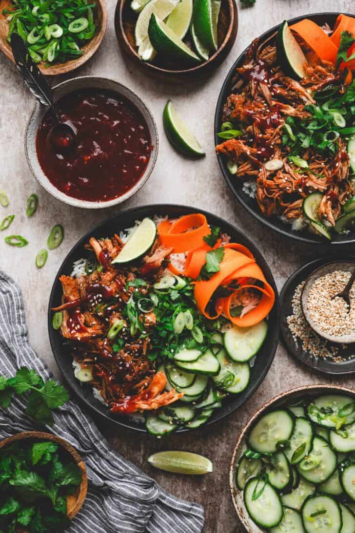 Asian style pulled pork rice bowl surrounded by cucumber salad, lime, cilantro, sesame seeds and extra sauce