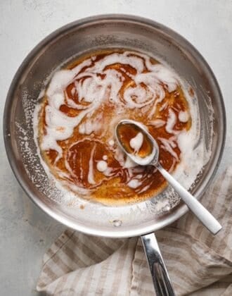 Overhead shot of a brown butter recipe in a stainless steel skillet, with deep amber milk solids swirled through the foam and a spoon resting in the pan