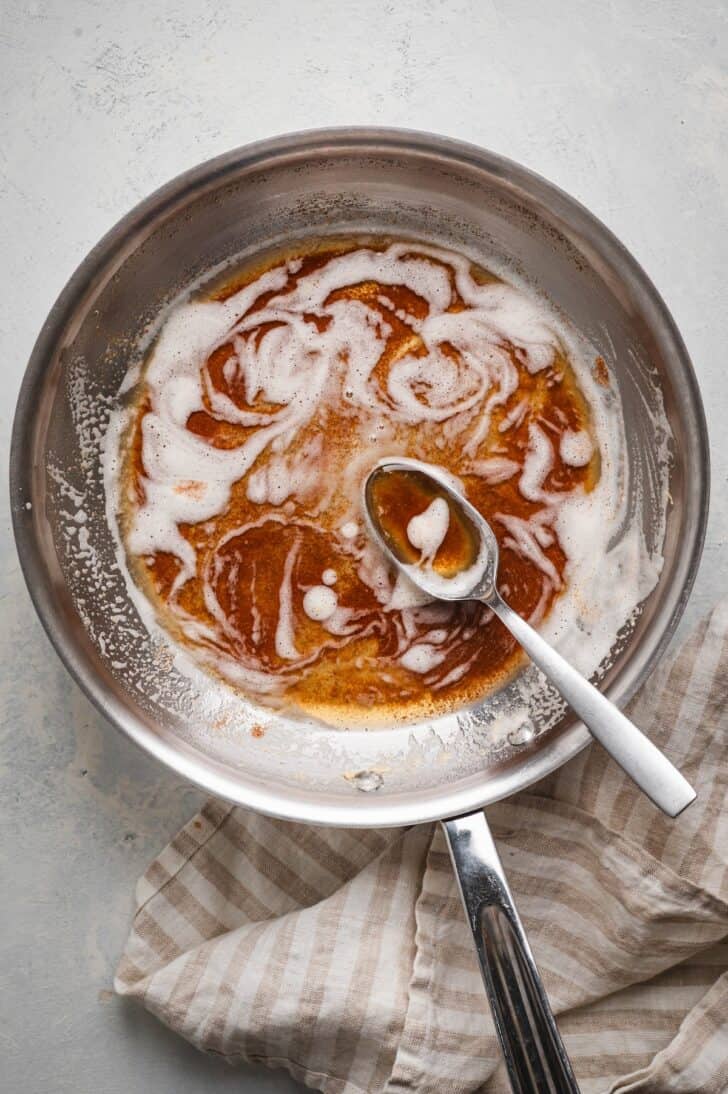 Overhead shot of a brown butter recipe in a stainless steel skillet, with deep amber milk solids swirled through the foam and a spoon resting in the pan
