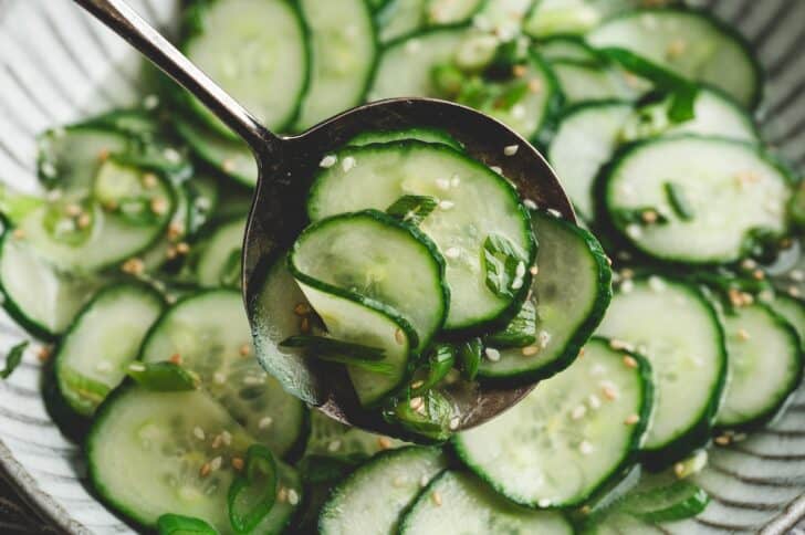 Close-up of a spoon lifting cucumber slices from a cucumber salad recipe asian style, glistening with dressing and flecked with sesame seeds