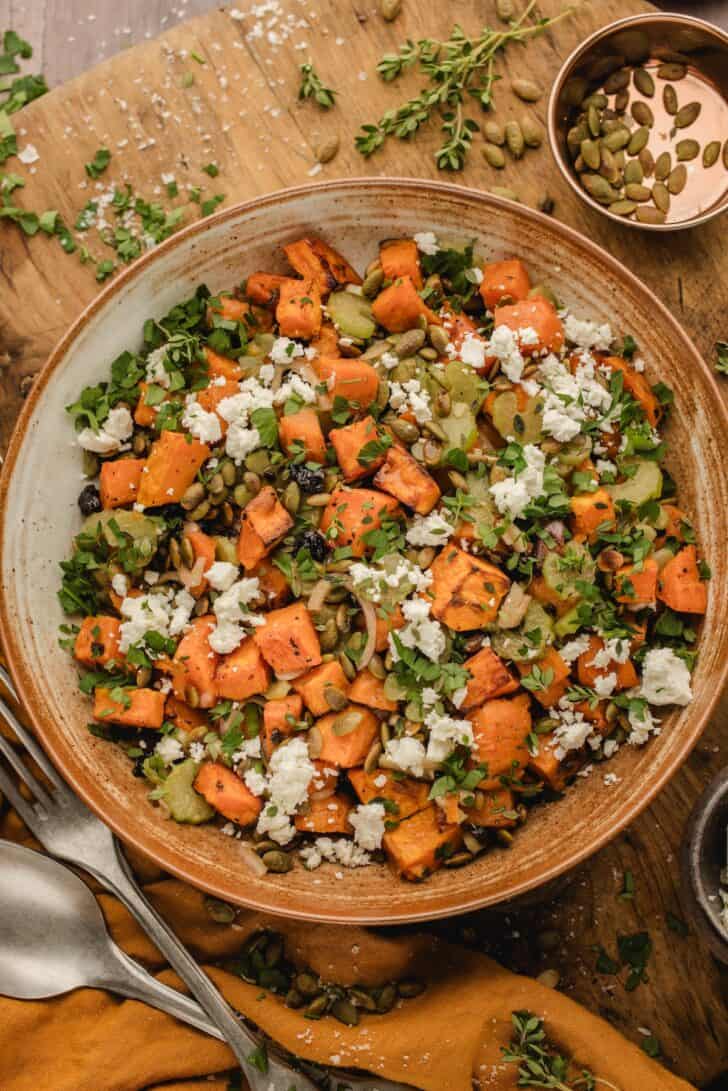 Overhead view of roasted sweet potato salad in a wide bowl, topped with crumbled feta, pepitas, and fresh herbs