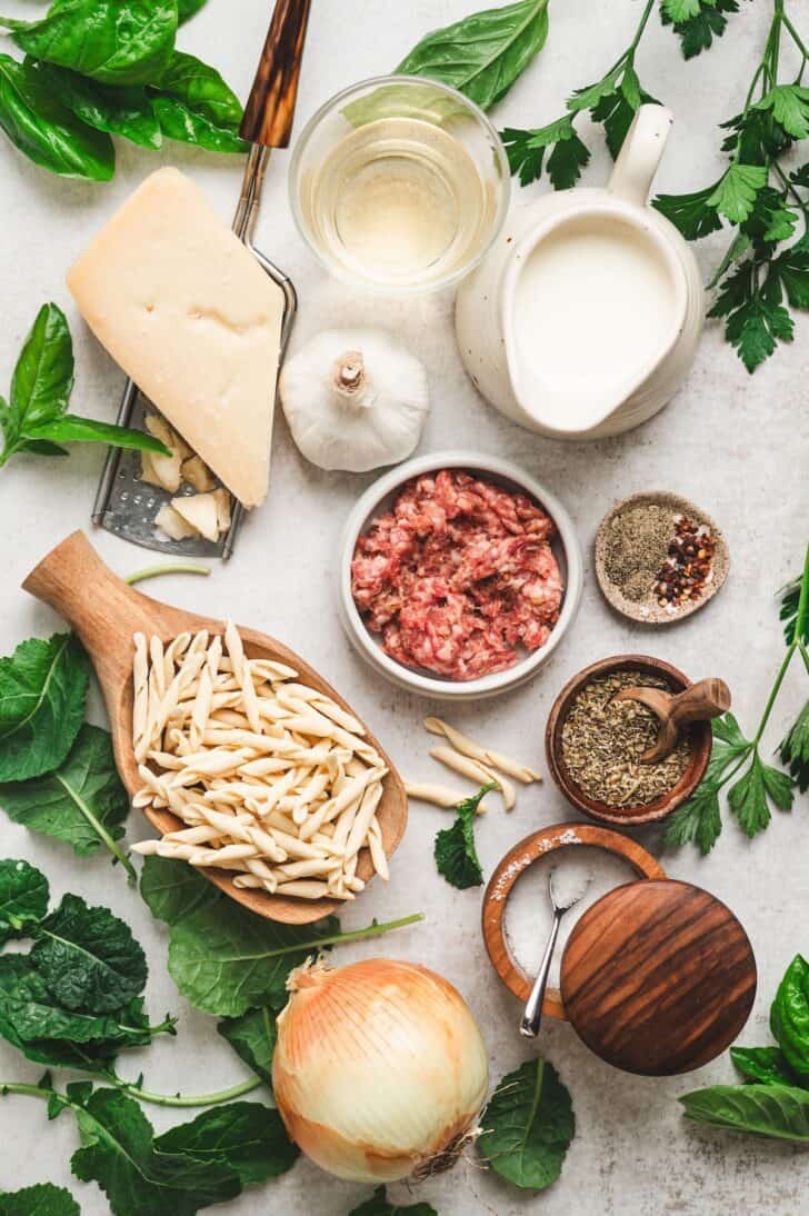Ingredients for italian sausage pasta laid out, including ground Italian sausage, pasta, Parmesan cheese, garlic, onion, heavy cream, white wine, and Italian seasoning