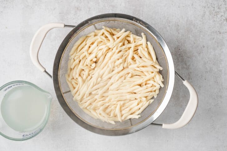 Process shot of cooked pasta draining in a colander next to a measuring cup of reserved pasta water