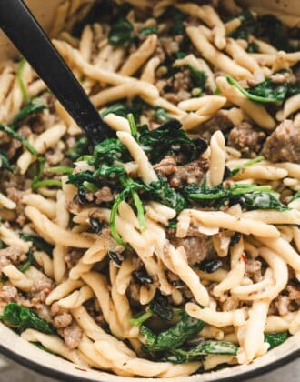 Scoop lifting a serving of pasta with crumbled Italian sausage, wilted greens, and cracked pepper from the pot, a favorite among italian sausage recipes