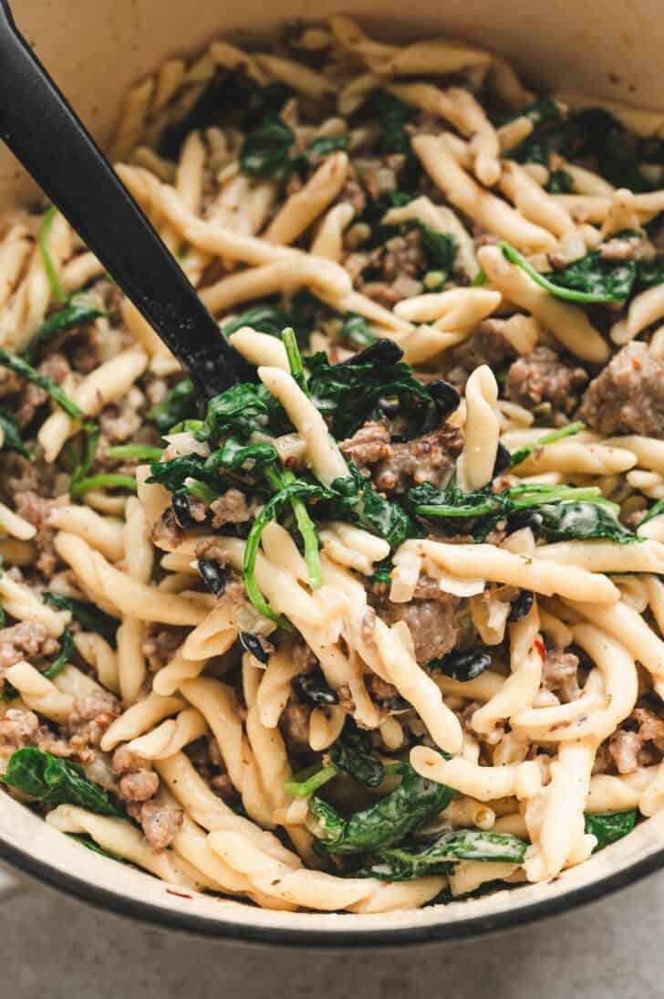 Scoop lifting a serving of pasta with crumbled Italian sausage, wilted greens, and cracked pepper from the pot, a favorite among italian sausage recipes