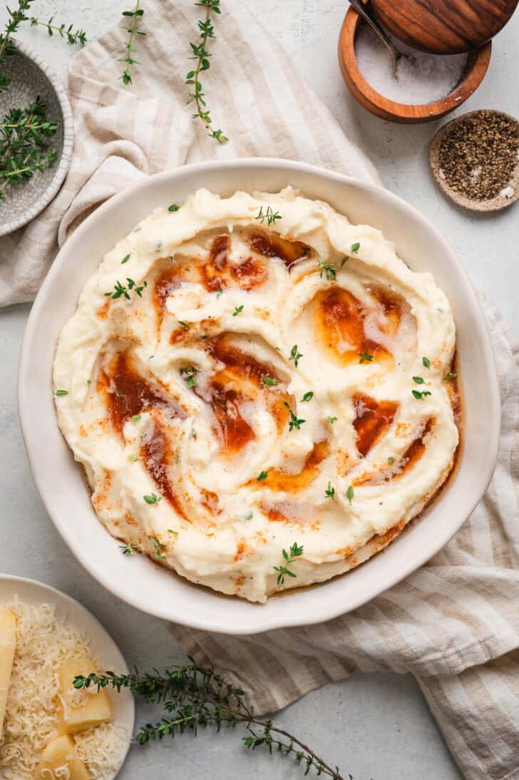 Overhead view of a bowl of parsnip puree swirled with warm brown butter and fresh thyme, surrounded by salt, pepper, and grated Parmesan
