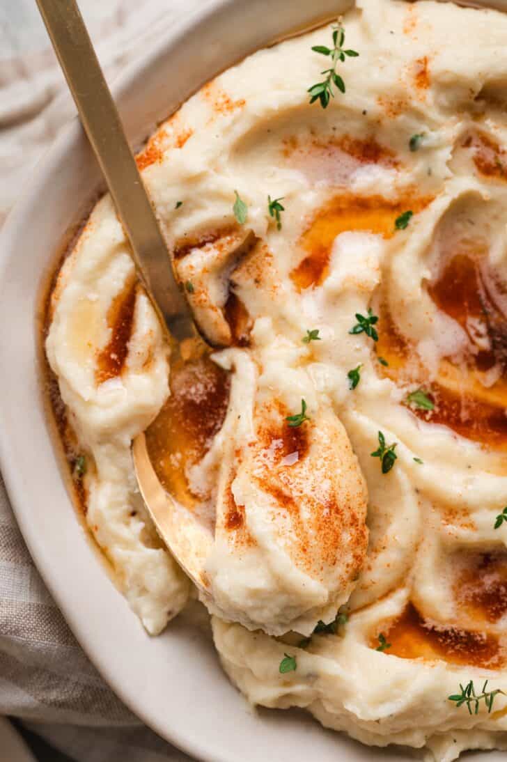 Close-up of a spoon digging into a bowl of mashed parsnips topped with brown butter and fresh thyme leaves
