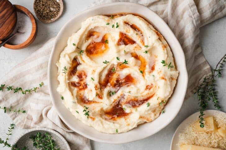 Overhead shot of a parsnip puree recipe in a shallow bowl, drizzled with brown butter and thyme, styled with a striped linen and bowl of Parmesan