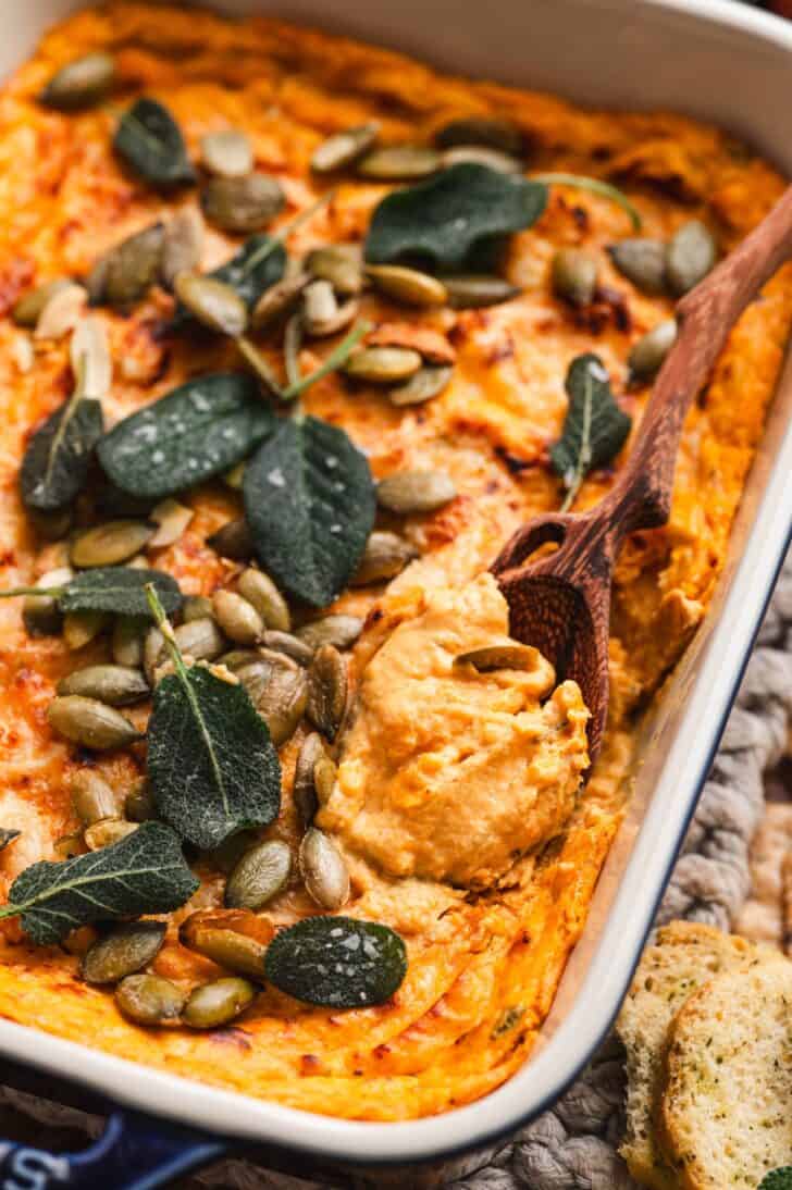 Close-up of a baked pumpkin dip recipe topped with crispy sage leaves and pepitas, with a spoon digging in and crostini alongside