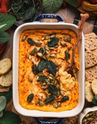 Overhead shot of pumpkin cream cheese dip topped with crispy sage and pepitas, surrounded by crackers and fall-toned linens