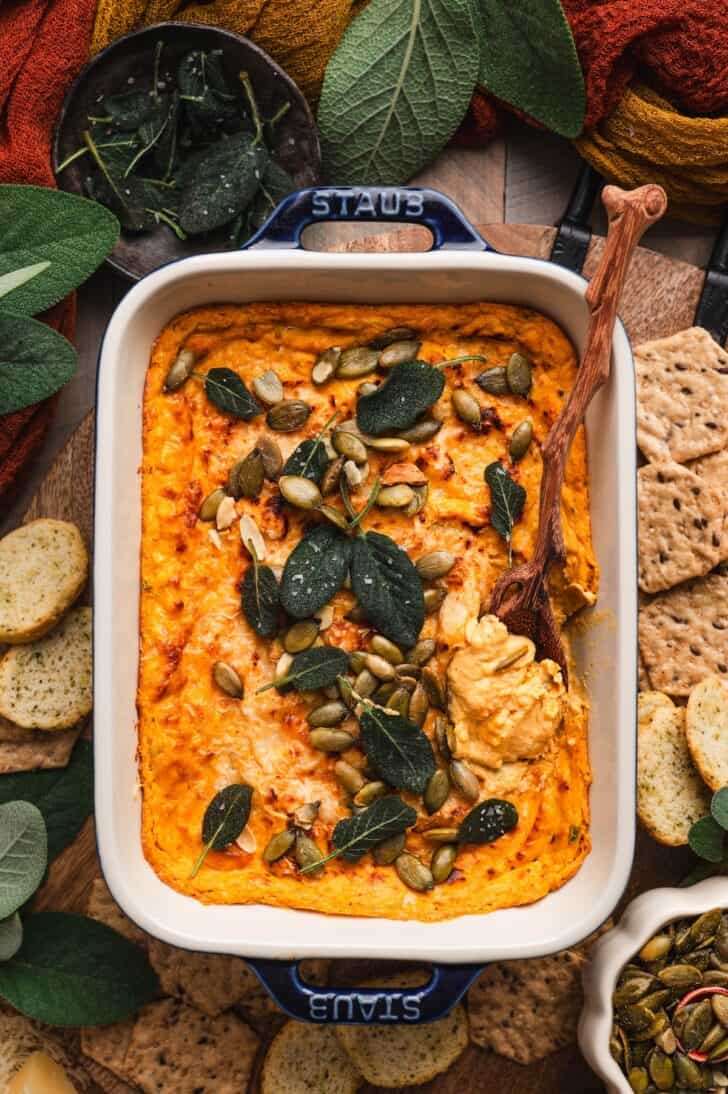 Overhead shot of pumpkin cream cheese dip topped with crispy sage and pepitas, surrounded by crackers and fall-toned linens