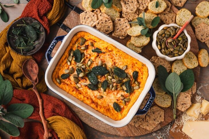 Wide spread of pumpkin dip with cream cheese served in a baking dish, surrounded by crackers, a wedge of Parmesan, and crispy sage leaves