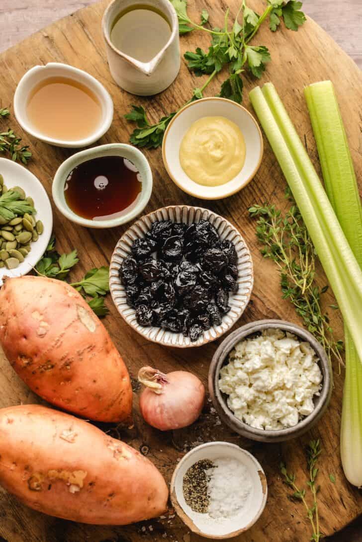 Ingredients laid out on a wood board, including whole sweet potatoes, celery, a shallot, dried cherries, feta cheese, pumpkin seeds, Dijon mustard, maple syrup, apple cider vinegar, and fresh herbs