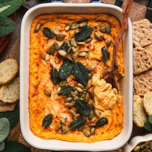 Overhead shot of pumpkin cream cheese dip topped with crispy sage and pepitas, surrounded by crackers and fall-toned linens