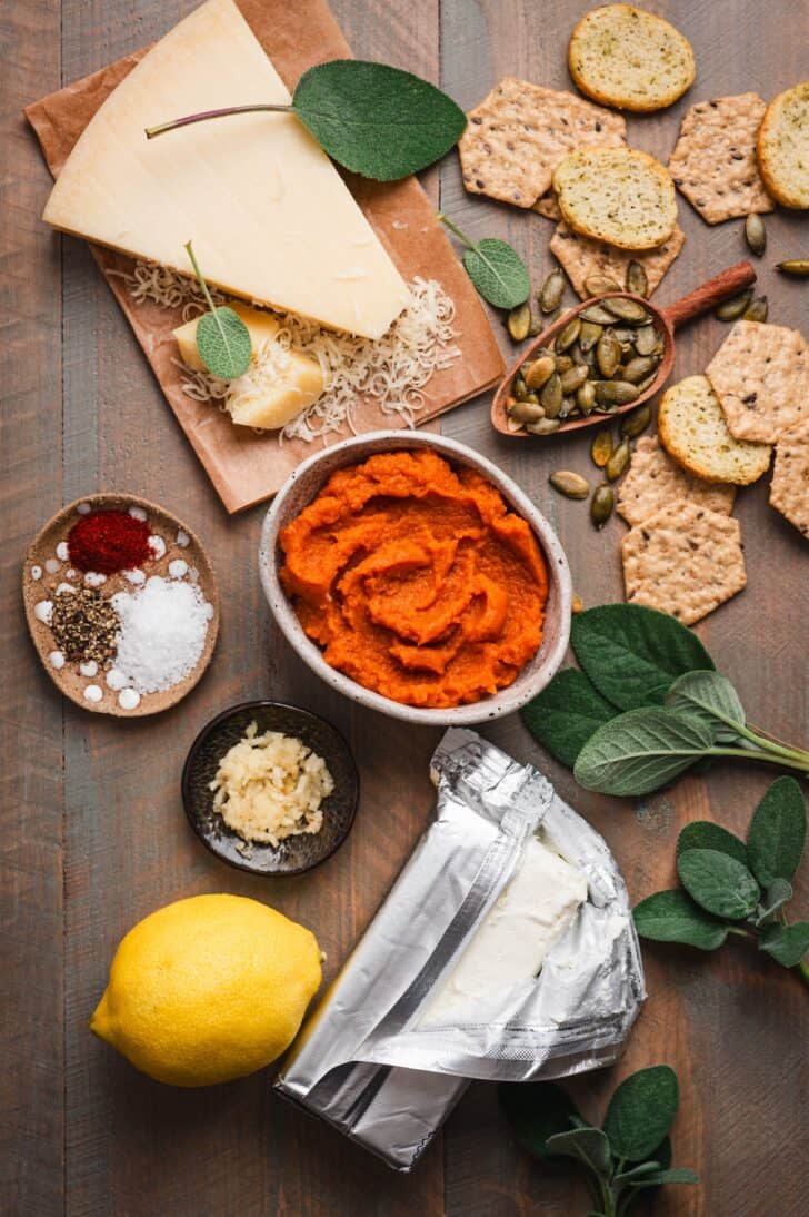 ngredients for the dip laid out, including a wedge of Parmesan cheese, crackers, pumpkin puree, spices, grated garlic, a lemon, foil-wrapped cream cheese, and fresh sage sprigs