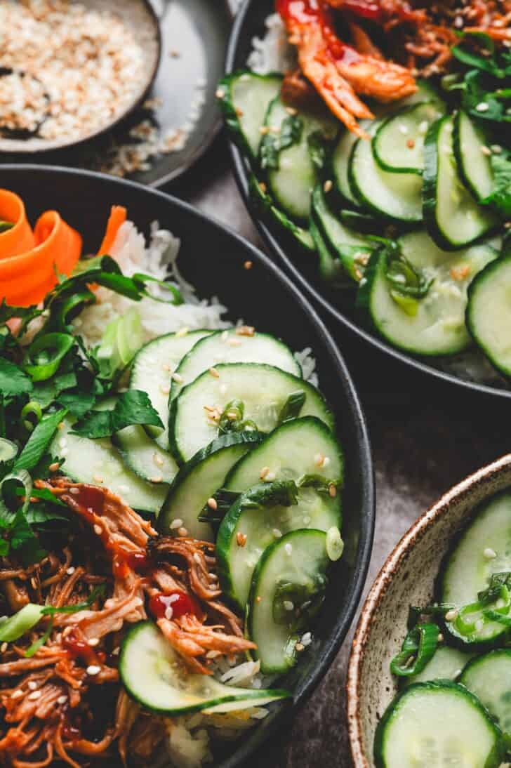 Sesame cucumber salad served on top of rice bowls with shredded pulled chicken, carrots, cilantro, and toasted sesame seeds