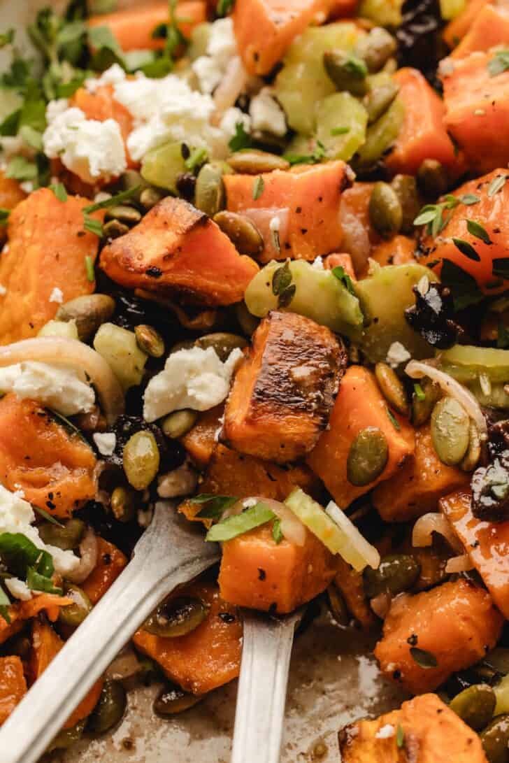 Extreme close-up of feta sweet potato salad with two forks lifting a bite, showing roasted sweet potato cubes, celery, and dried cherries