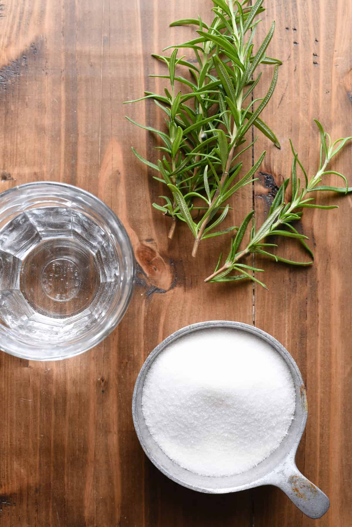 Overhead photo of cup of sugar, glass of water and rosemary sprigs on wooden table.